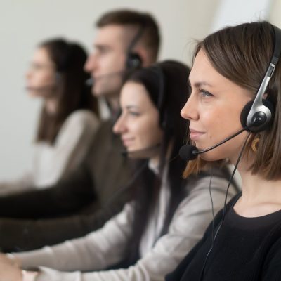 pexels-photo-7035859-7035859 Side view of young coworkers in casual clothes sitting in light workplace with headphones with microphone while surfing on laptop